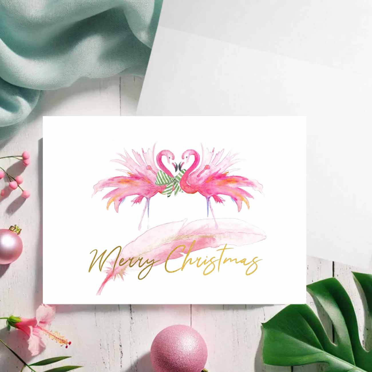 flat lay of watercolor pink flamingo Christmas cards with the words Merry Christmas in gold foil. Unique art and design by Victoria Grigaliunas of Do Tell A Belle a woman owned small business in Southern Florida. The card is blank inside.
