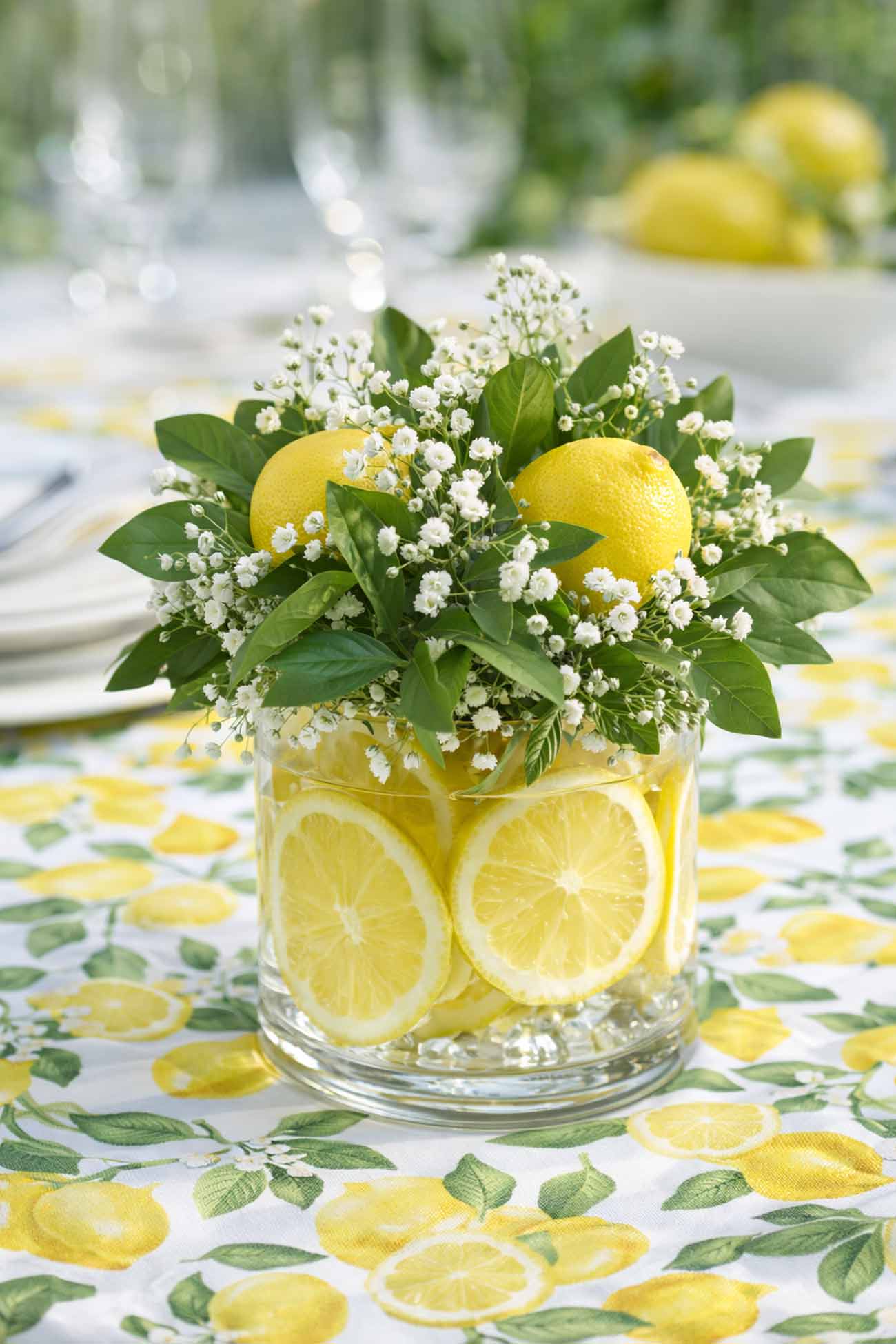 lemon bridal shower centerpiece with lemon slices in a glass vase and greenery on a yellow lemon tablecloth
