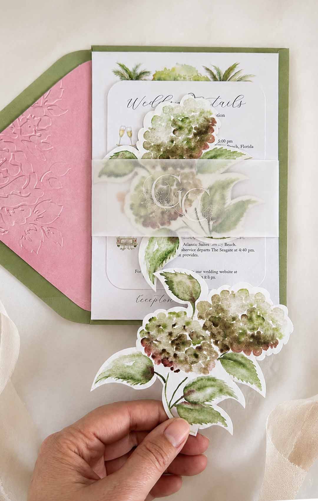 ALT TEXT Watercolor wedding invitations featuring olive green envelopes, a pink embossed envelope liner, vellum monogram belly band, and a die cut hydrangea RSVP card in a custom wedding stationery suite.