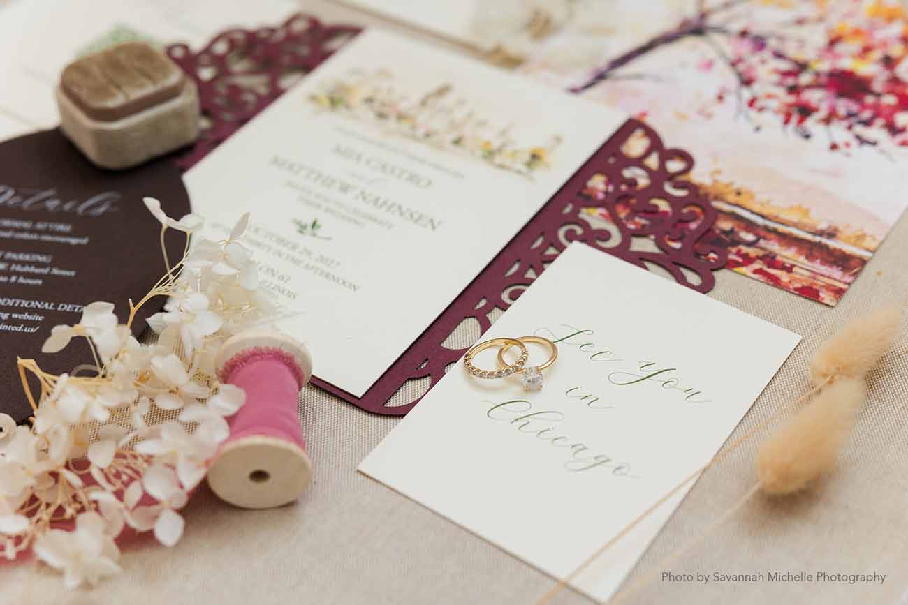 Elegant watercolor wedding invitations featuring a burgundy laser-cut gatefold, floral watercolor artwork, and coordinating details card, styled with wedding rings and romantic accessories.