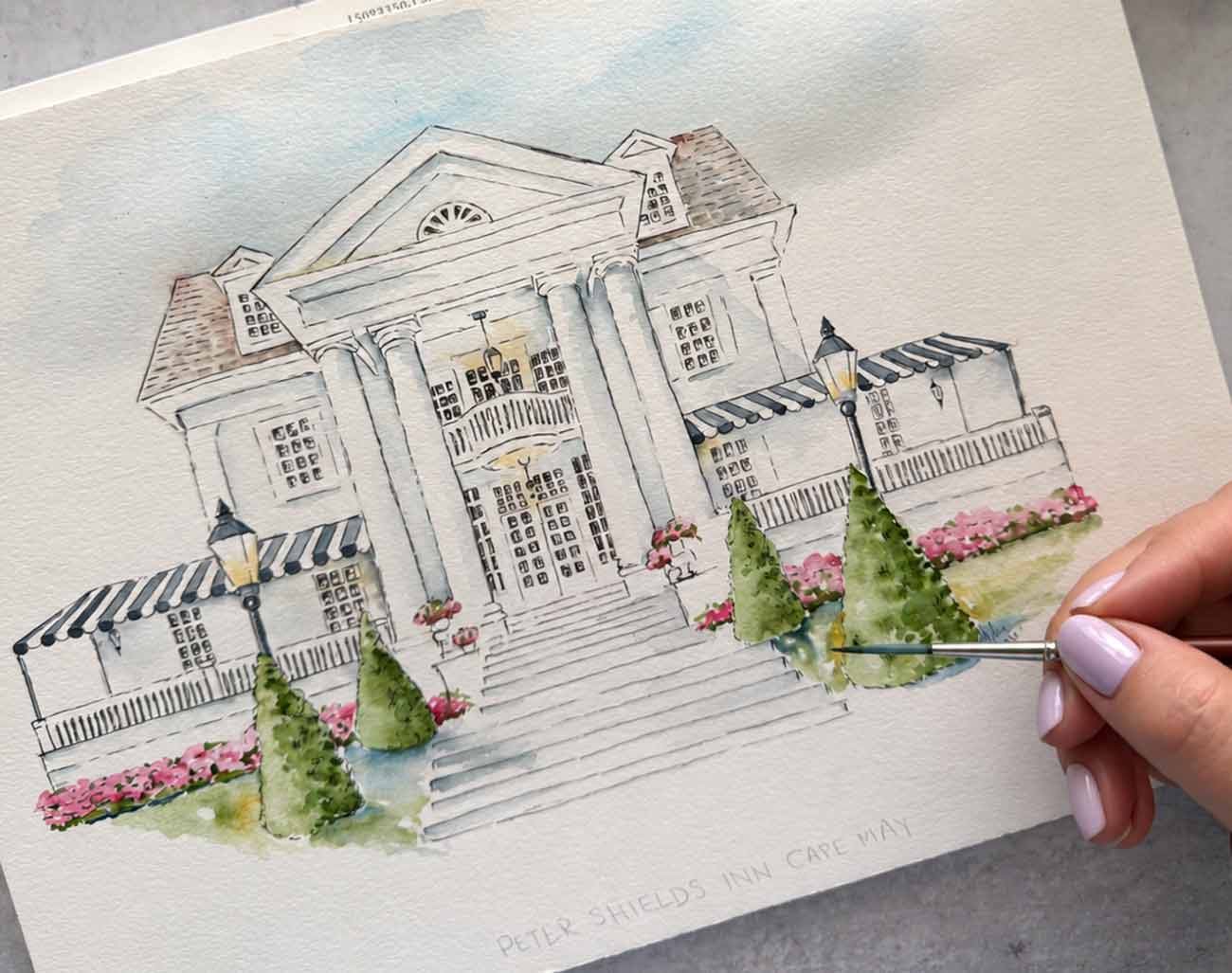Watercolor wedding venue artist Victoria Grigaliunas hand-painting an architectural illustration of Peter Shields Inn in Cape May, featuring elegant columns, pink flowers, and lush greenery.